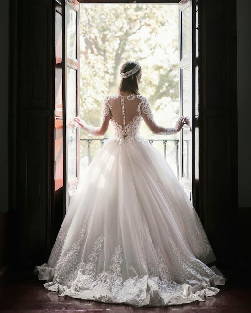 A bride in a lace gown looking out through open balcony doors